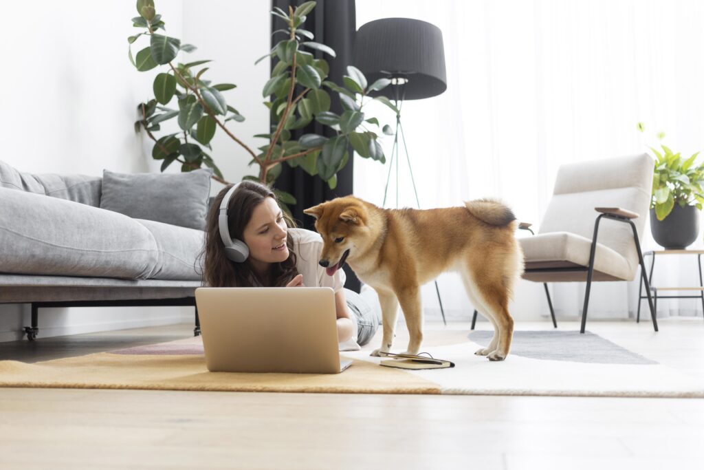 Woman wearing headphones lies on the floor using a laptop while her Shiba Inu stands beside her in a bright, modern living room—a perfect example of the comfort and joy that comes with renting with pets.