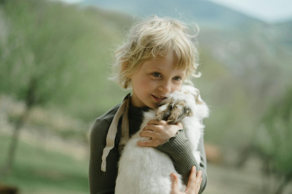 A young child with blonde hair holds a small white goat, showcasing pets for children, outdoors in a natural green setting.