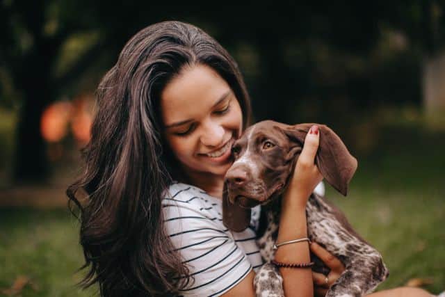A woman with long dark hair, wearing a striped shirt, smiles while holding her new puppy outdoors, reflecting the joy and responsibility of preventive health care for dogs.