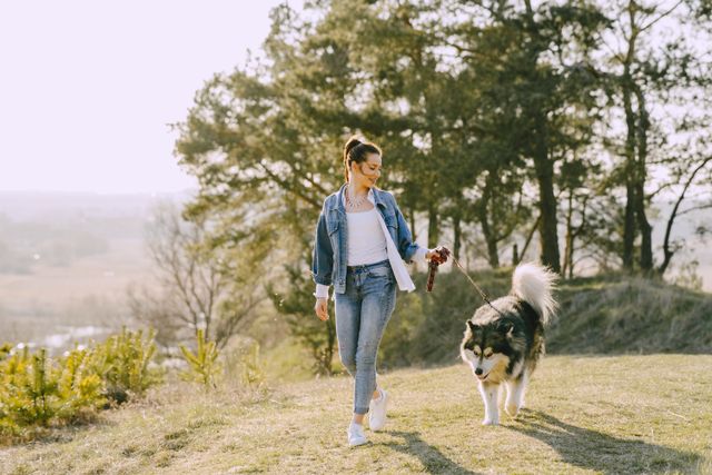 A woman in casual clothes walks a large dog on a leash through a grassy area with trees, enjoying the sunny day as she discusses routine pet care.