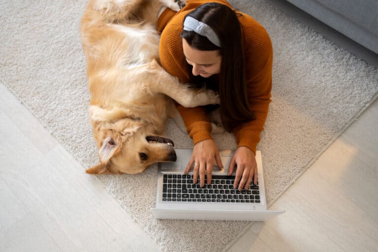A woman lies on a carpet using her laptop and exploring an AI pet app, while her golden retriever playfully rests its paw on her arm. Embrace the connection between pet technology and quality bonding moments.