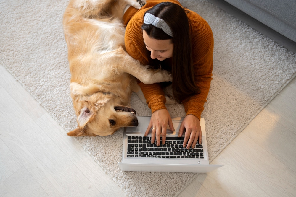 A woman lies on a carpet using her laptop and exploring an AI pet app, while her golden retriever playfully rests its paw on her arm. Embrace the connection between pet technology and quality bonding moments.