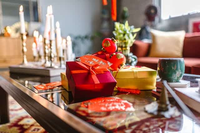Festive gifts wrapped in red and yellow paper with ribbons sit on a living room table, evoking the spirit of Chinese New Year. Candles, fruit, and a green mug create a warm ambiance to welcome the Year of the Snake.
