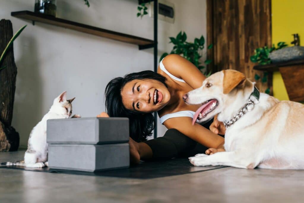 A woman smiles while stretching on a yoga mat indoors, accompanied by a dog and a cat sitting nearby, ensuring the space is safe by avoiding anything harmful to pets like citronella candles.