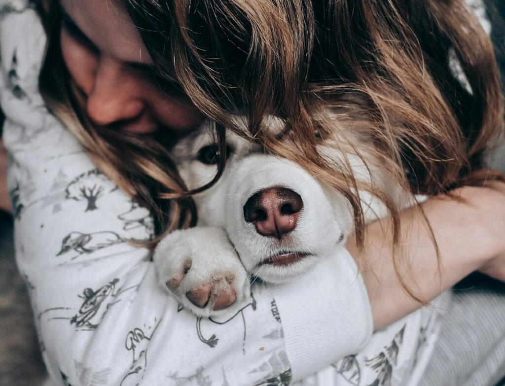 A person with long hair hugs a white dog closely, part of their hair veiling the dog's face, capturing a poignant moment as they grieve the loss of a pet.