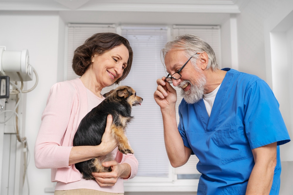 A woman holding a small dog discusses pet claims with a male veterinarian in blue scrubs inside a veterinary clinic.