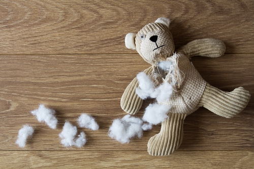 A stuffed teddy bear with a torn seam and loose stuffing sits on the wooden floor, posing a risk of accidental ingestion due to foreign objects potentially hidden inside.