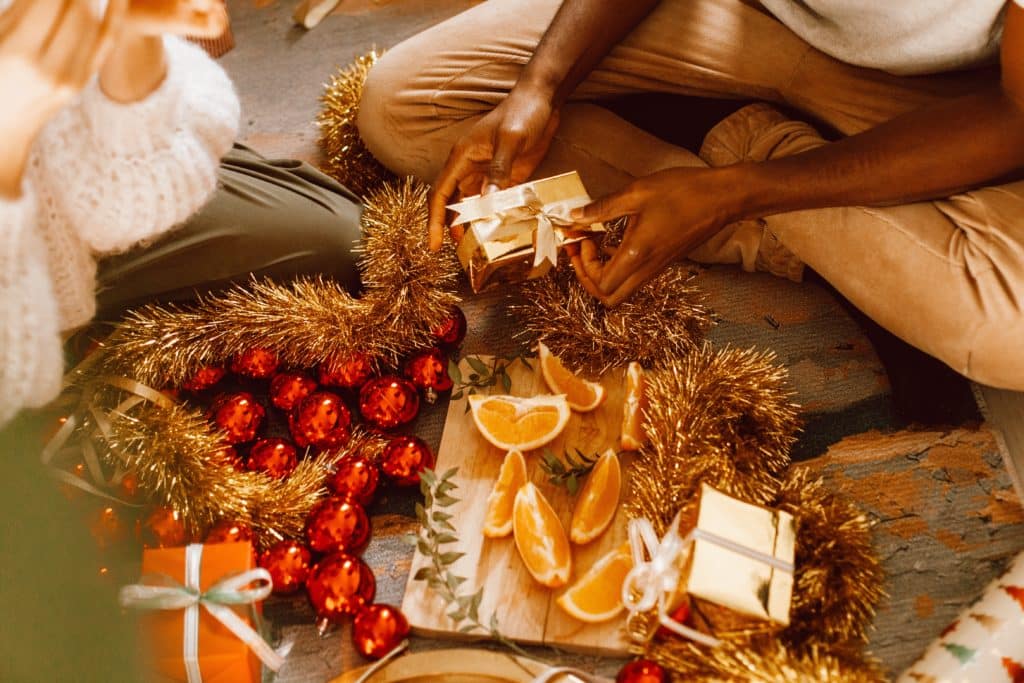 People sitting on the floor, surrounded by gold tinsel, red ornaments, and small gift boxes, enjoy sliced oranges on a wooden board.