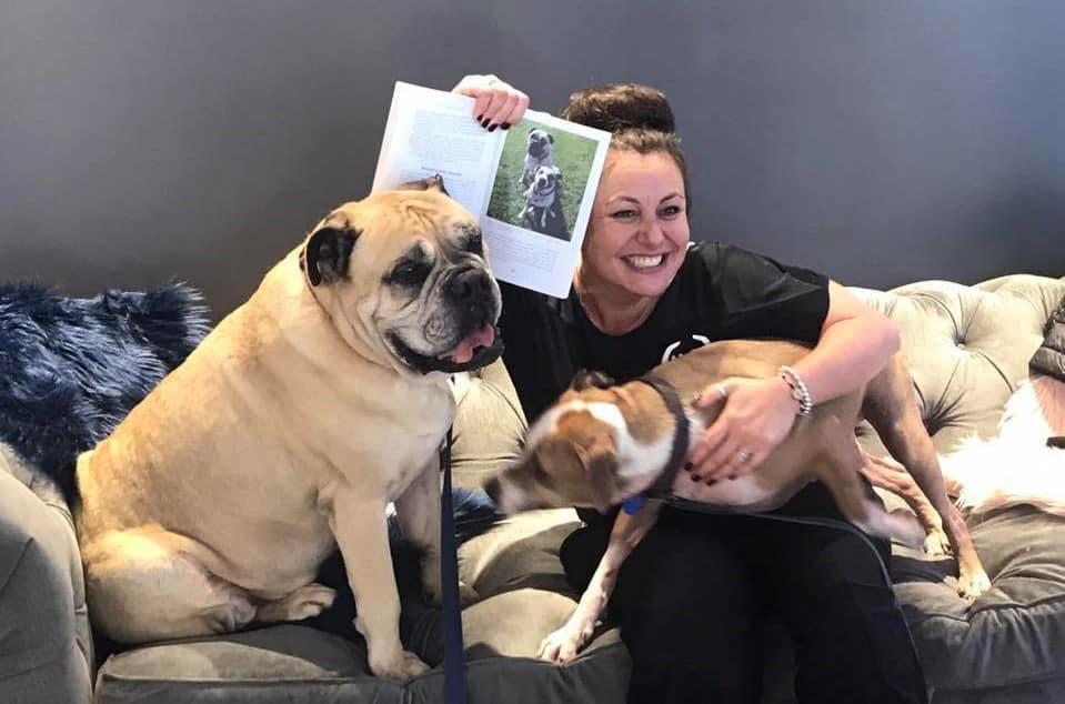 Canine Emotional Detox dog trainer Maria Alojaman holds up a book featuring her dogs (which sit alongside her)