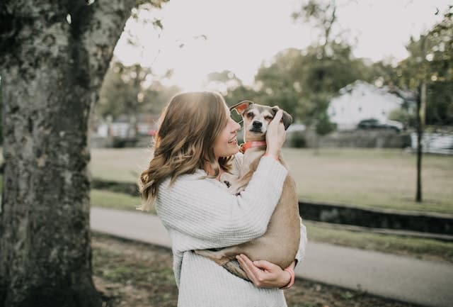 Smiling Woman Carrying Dog Near Tree. Whether you're a dog mom or a cat mom, here's how to enjoy Mother's Day