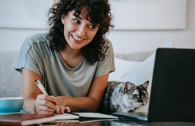 Glad woman with cat writing in planner while using laptop. Whether you're a dog mom or a cat mom, here's how to enjoy Mother's Day