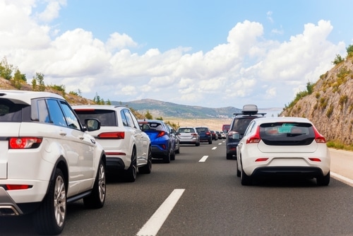 Cars are lined up in heavy traffic on a highway surrounded by hills under a partly cloudy sky, as drivers stay alert for stray animals that may unexpectedly cross the road.