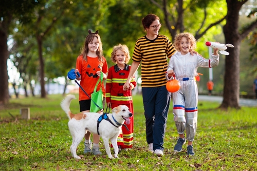A group of kids in costumes and an adult walk a pup in a park. 