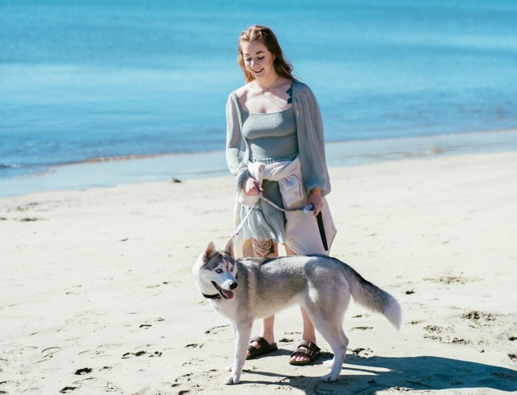 A woman standing on the beach with a husky dog, holding a dog leash.