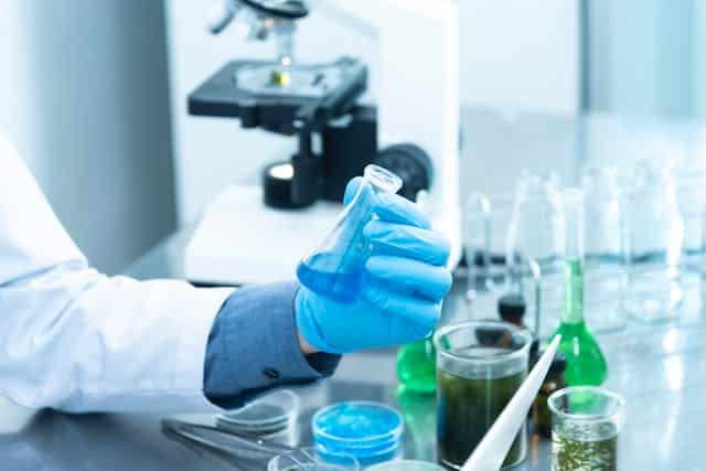 In front of a microscope, a scientist diligently holds a flask of liquid while conducting an experiment.