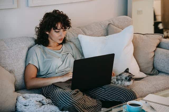 A woman enjoying her laptop on the couch with a cat beside her, occasionally taking breaks to play cat games or indulge in a round of the Exploding Kittens card game with housemates.
