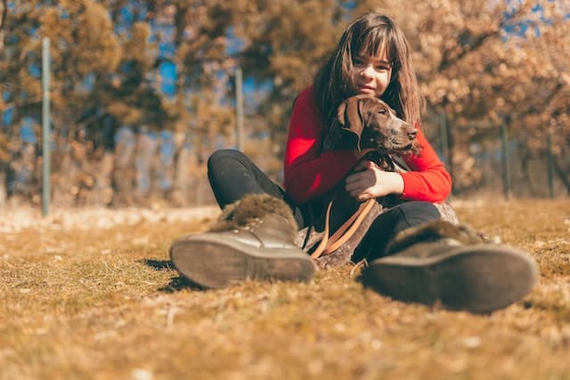 A girl is sitting on the ground with her dog who is protected by pet accident insurance.