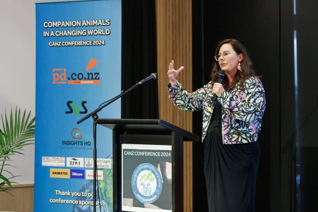 A woman presenting at the "Companion Animals NZ conference 2024," standing behind a lectern with a microphone, gesturing with her right hand.