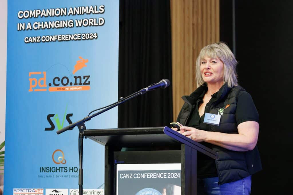 A woman speaking at a lectern with a "Companion Animals NZ (CANZ) conference 2024" banner in the background.