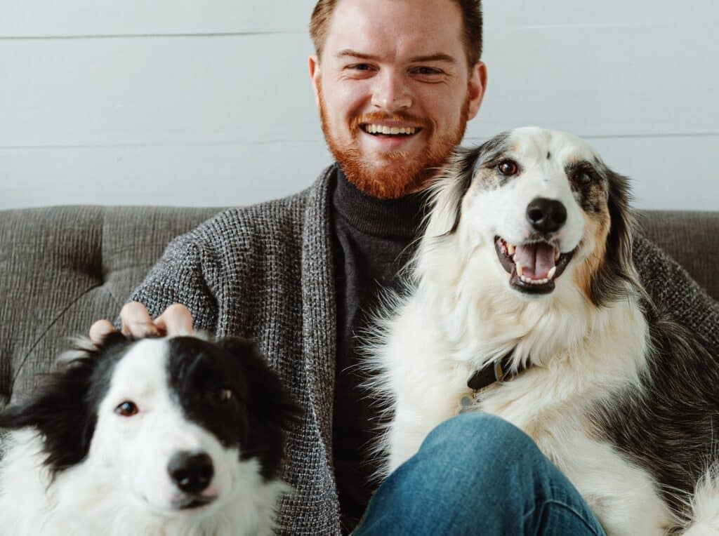 A smiling man with a red beard sitting on a couch, flanked by two happy border collies displaying plaque on their teeth (which will be brushed away later).