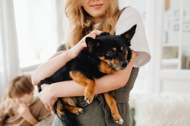 A person is holding a small black and brown dog, showcasing the joy of sustainable pet ownership. Another individual is seated in the background, looking down. The setting appears to be a comfortable indoor environment.