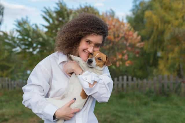 A smiling young woman with curly hair celebrating International Dog Day by holding a small dog in a lush outdoor setting with autumn trees in the background.