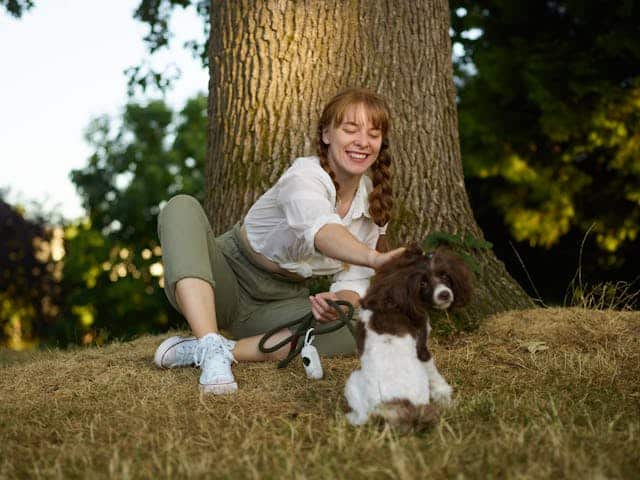 A woman sitting by a tree, laughing, as she plays with her brown and white dog in a sunlit park during International Dog Day.