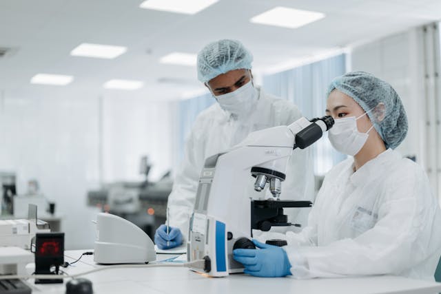 Two scientists in protective gear work in a laboratory, one looking through a microscope while the other observes, discussing how AI can help pets through advancements in data science.