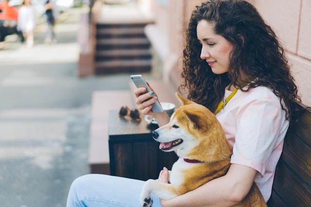 A woman with curly hair sits on a bench, holding a dog in her lap while looking at her smartphone. The bench is outside, with blurred background elements. She appears to be exploring an AI pet app designed to enhance the well-being of pets through data-driven insights.