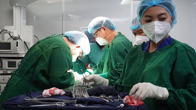 Medical team dressed in green surgical attire and masks performing surgery in an operating room, with a focus on a nurse preparing surgical instruments, reminiscent of the meticulous care given to keep a dog's cast dry after treating an infected dog wound.