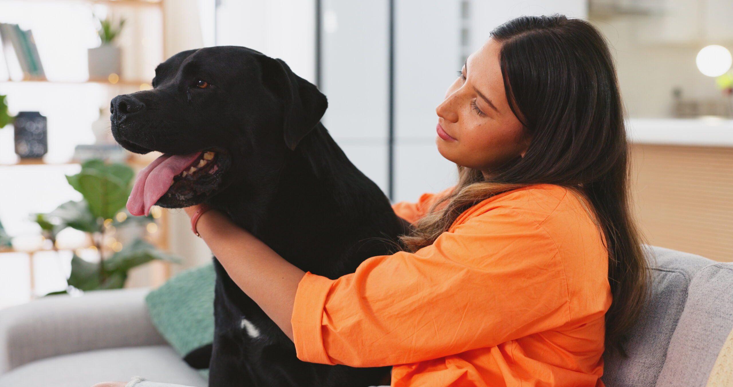 A woman in an orange shirt sits on a couch, hugging and smiling at a large black dog with its tongue out, grateful for the relief Cytopoint for dogs has brought to her pet's atopic dermatitis.
