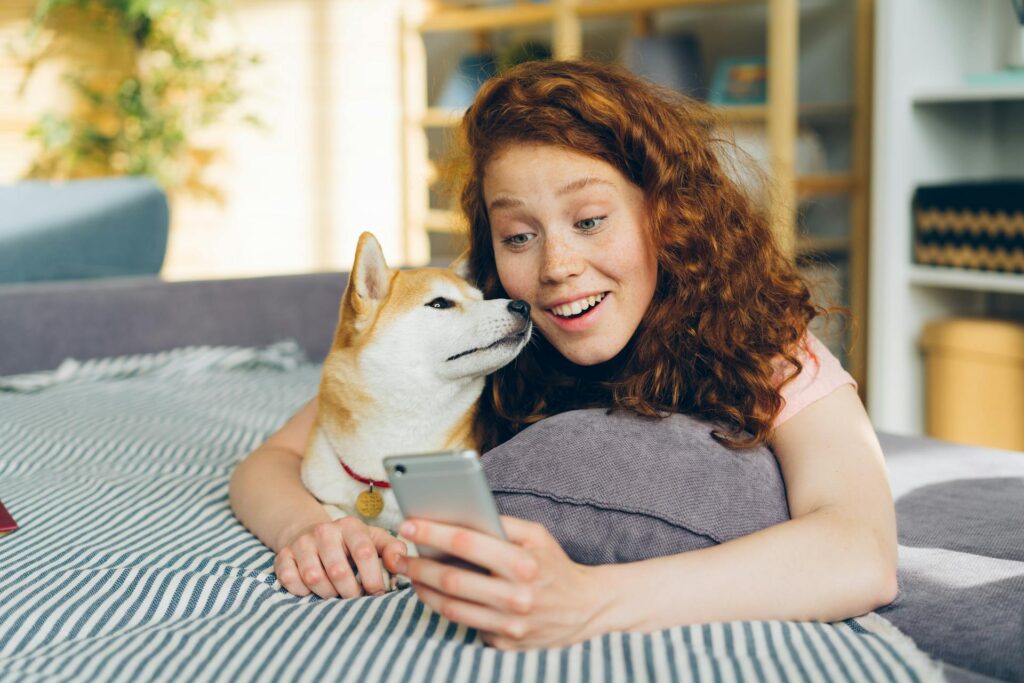 A person with red curly hair lies on a striped blanket next to a Shiba Inu dog, both looking at a smartphone screen, perhaps exploring the best AI apps together.