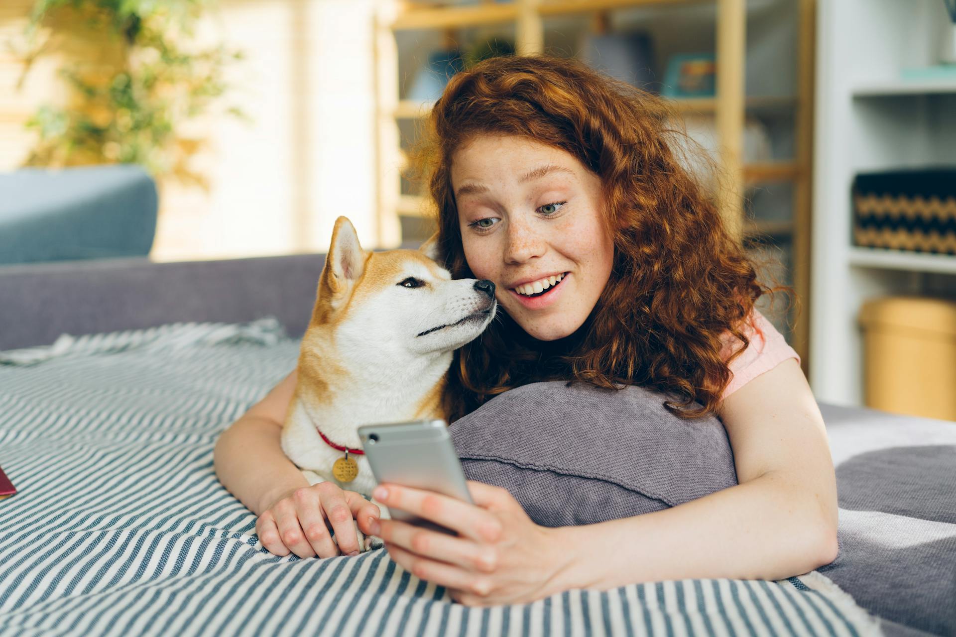A person with red curly hair lies on a striped blanket next to a Shiba Inu dog, both looking at a smartphone screen, perhaps exploring the best AI apps together.