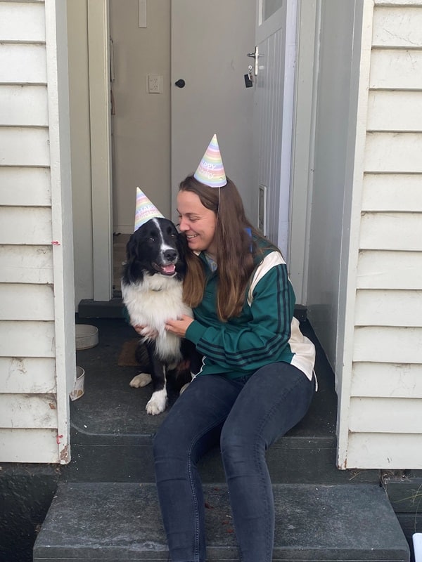 A woman and Nelson the Border Collie, both wearing birthday hats, sit on the steps of a white house. The woman is smiling and looking at the dog.