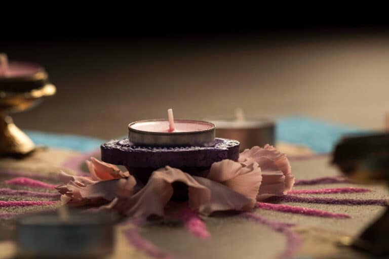 A pink tea light candle in a decorative holder surrounded by flower petals and colored sand for Diwali 2024.