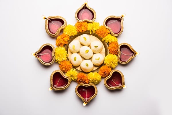 A circular arrangement of lit diya lamps and yellow-orange flowers frames a plate of round, white sweets with green garnish on a white background, evoking the vibrant spirit of Diwali 2024.