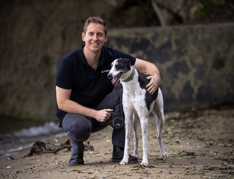 A man kneels on a sandy beach next to his black and white dog, holding its leash. With holistic approach, he follows vet advice to soothe his dog's itchy skin. In the background, a stone wall and greenery complete the serene setting.