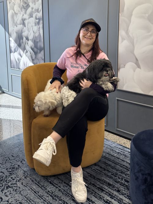 Woman sitting on a tan chair holding two small dogs, one black and one white, in a modern room with patterned walls, celebrating Pink Shirt Day to support anti bullying day NZ.