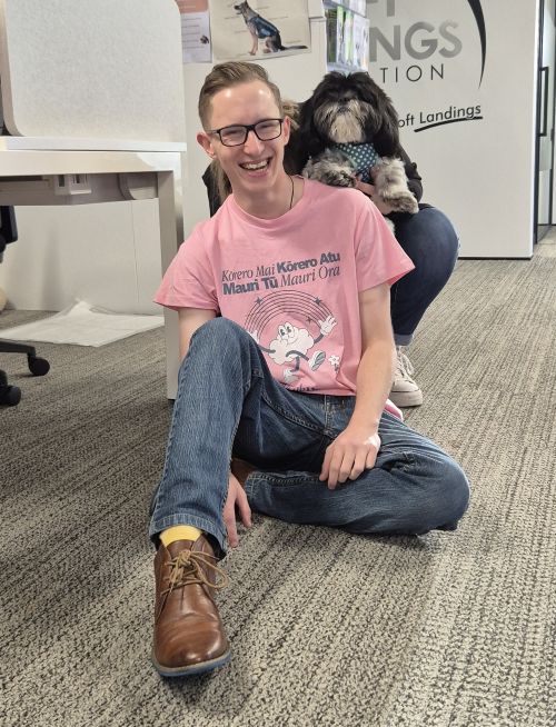 A person in a pink T-shirt and jeans sits on the floor, smiling, with a small black and white dog on their shoulders in an office setting, celebrating Pink Shirt Day and supporting anti bullying day NZ.