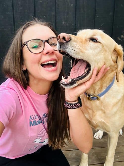 A woman wearing glasses and a pink shirt smiles while holding a yellow dog’s face as the dog opens its mouth near her cheek, celebrating Pink Shirt Day—an event that proudly supports anti bullying day NZ.