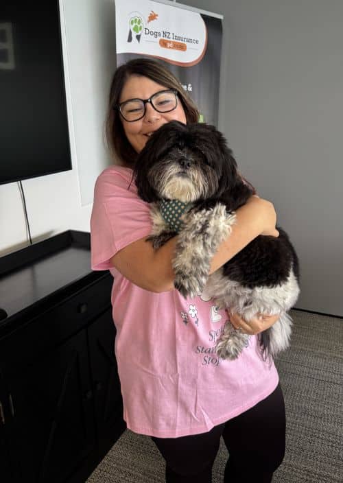 A woman wearing glasses and a pink t-shirt holds a black and white dog in her arms, standing indoors near a Dogs NZ Insurance banner, showing her support for Pink Shirt Day and anti bullying day NZ.