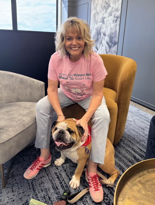 A woman in a pink T-shirt and jeans sits on a chair, smiling with her hands on a brown and white bulldog wearing a red bandana, celebrating Pink Shirt Day—an event promoting kindness for Anti Bullying Day NZ.
