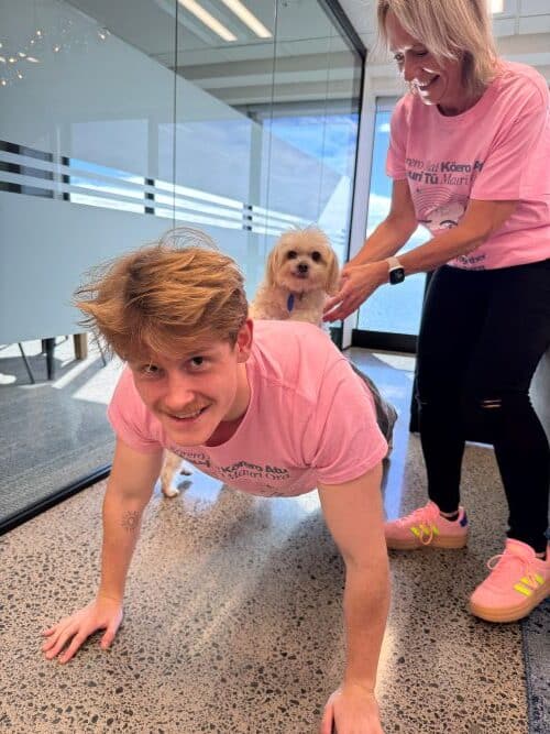 A young man does push-ups with a small dog on his back, while an older woman in pink athletic wear—celebrating Pink Shirt Day, anti bullying day NZ—steadies the dog in an indoor space.