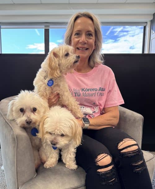 A woman sits on a gray chair indoors, smiling, with three small white dogs on her lap. She is wearing a pink T-shirt to celebrate Pink Shirt Day, pairing it with ripped black jeans. A window shows a blue sky outside.