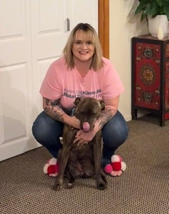 A woman in a pink shirt and slippers crouches behind a brown dog sitting on carpet, with the dog licking its nose—capturing the spirit of Pink Shirt Day and raising awareness for anti bullying day nz.