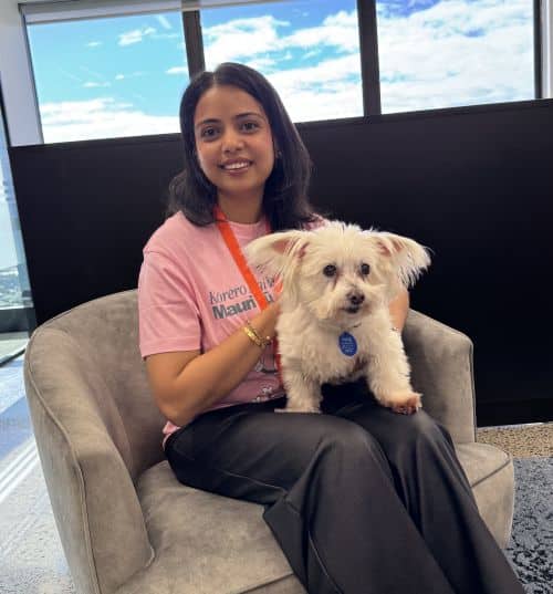 A woman sits in a gray chair holding a small white dog. She is wearing a pink shirt in support of Pink Shirt Day, highlighting anti bullying day NZ, with large windows and a blue sky in the background.