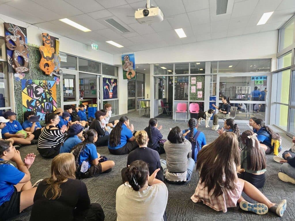 A group of students sit in a circle on the classroom floor, listening to a speaker and a friendly support dog promoting pawsitivity; colourful artwork and posters decorate the room.