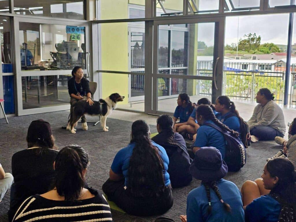 A group of students sits on the floor facing a woman and a support dog during an indoor presentation near large windows, learning about the power of pawsitivity.