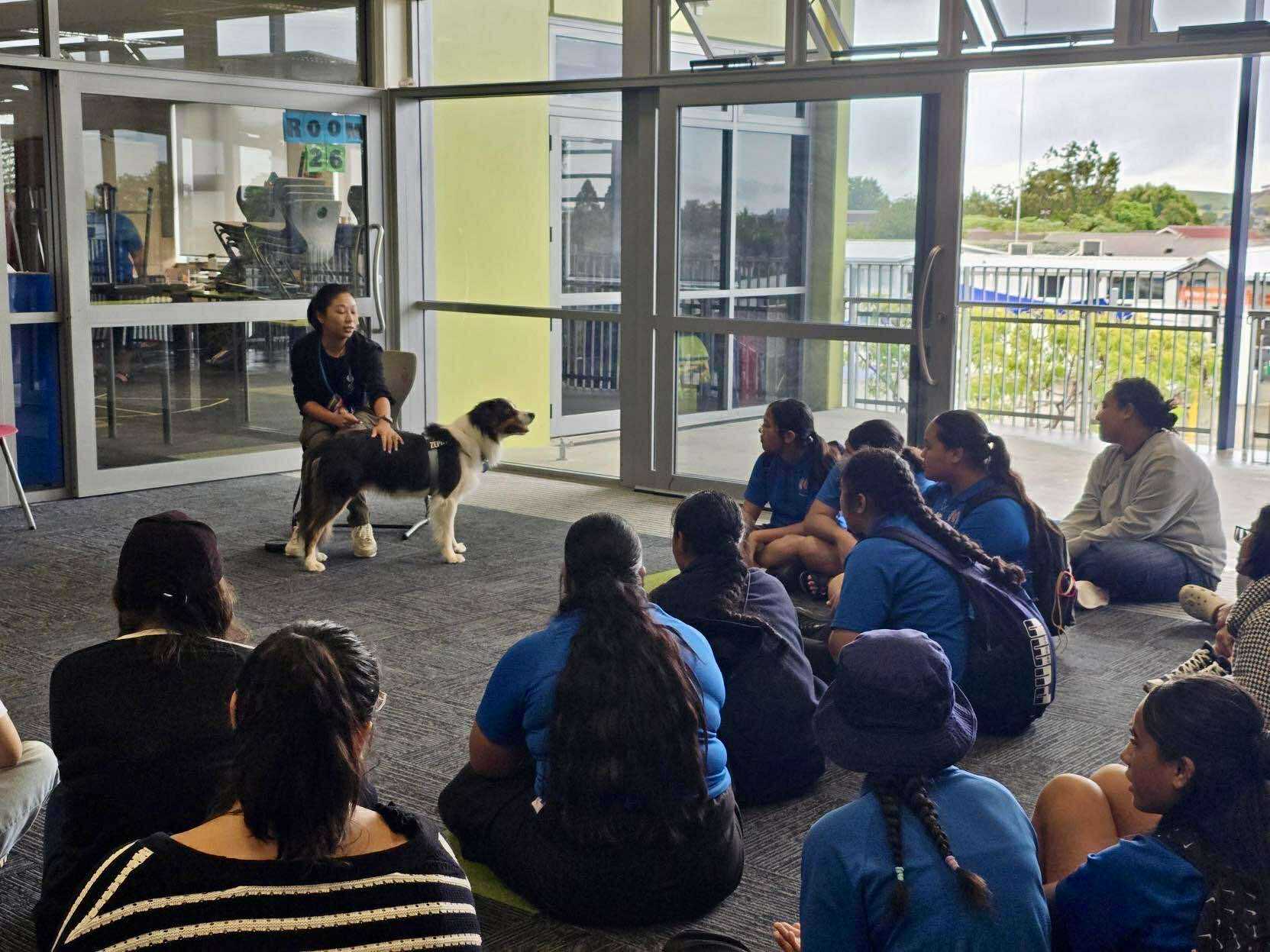 A group of students sits on the floor facing a woman and a support dog during an indoor presentation near large windows, learning about the power of pawsitivity.
