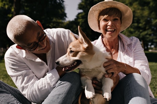 Two older adults sit on grass outdoors, smiling and petting a corgi dog. They are wearing light clothing and sun hats, enjoying time together while considering the cost of owning a dog. Trees and sunlight are visible in the background.